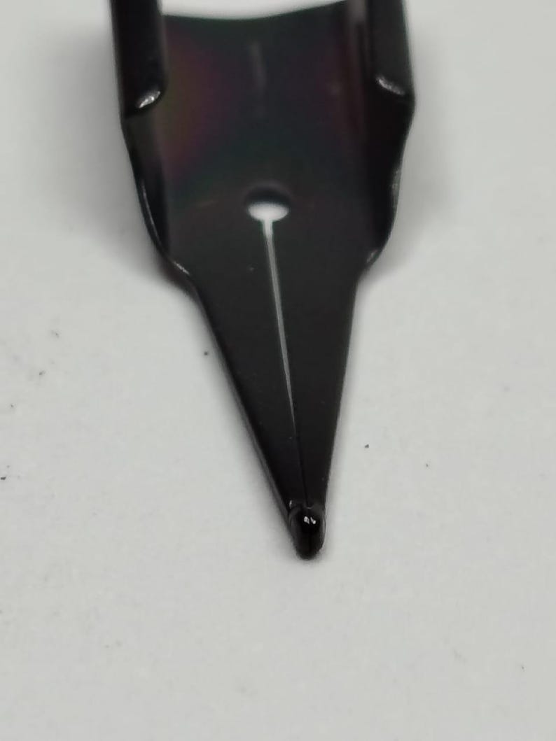 Puede incluir: Primer plano de una plumilla de pluma estilogr&aacute;fica negra. La plumilla tiene una punta fina con una ranura en el centro. La plumilla es de metal y tiene un acabado brillante. El fondo es una superficie blanca lisa.