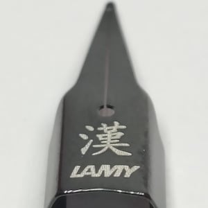 Puede incluir: Una plumilla de pluma estilogr&aacute;fica LAMY negra con el car&aacute;cter chino "漢" grabado. La plumilla tiene una punta afilada y un peque&ntilde;o agujero. El logotipo de LAMY est&aacute; debajo del car&aacute;cter.