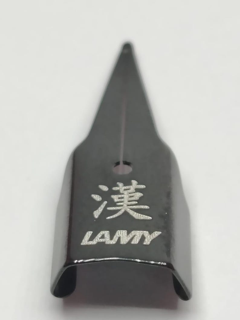 Puede incluir: Una plumilla de pluma estilogr&aacute;fica LAMY negra con el car&aacute;cter chino "漢" grabado. La plumilla tiene una punta afilada y un peque&ntilde;o agujero. El logotipo de LAMY est&aacute; debajo del car&aacute;cter.