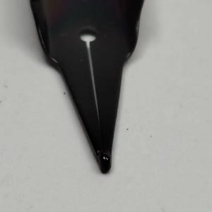 Puede incluir: Primer plano de un plum&iacute;n de pluma estilogr&aacute;fica negro. El plum&iacute;n es de metal y tiene una punta fina. Una fina ranura recorre el centro del plum&iacute;n y se ve un peque&ntilde;o agujero cerca de la parte superior. El plum&iacute;n est&aacute; sobre un fondo blanco.