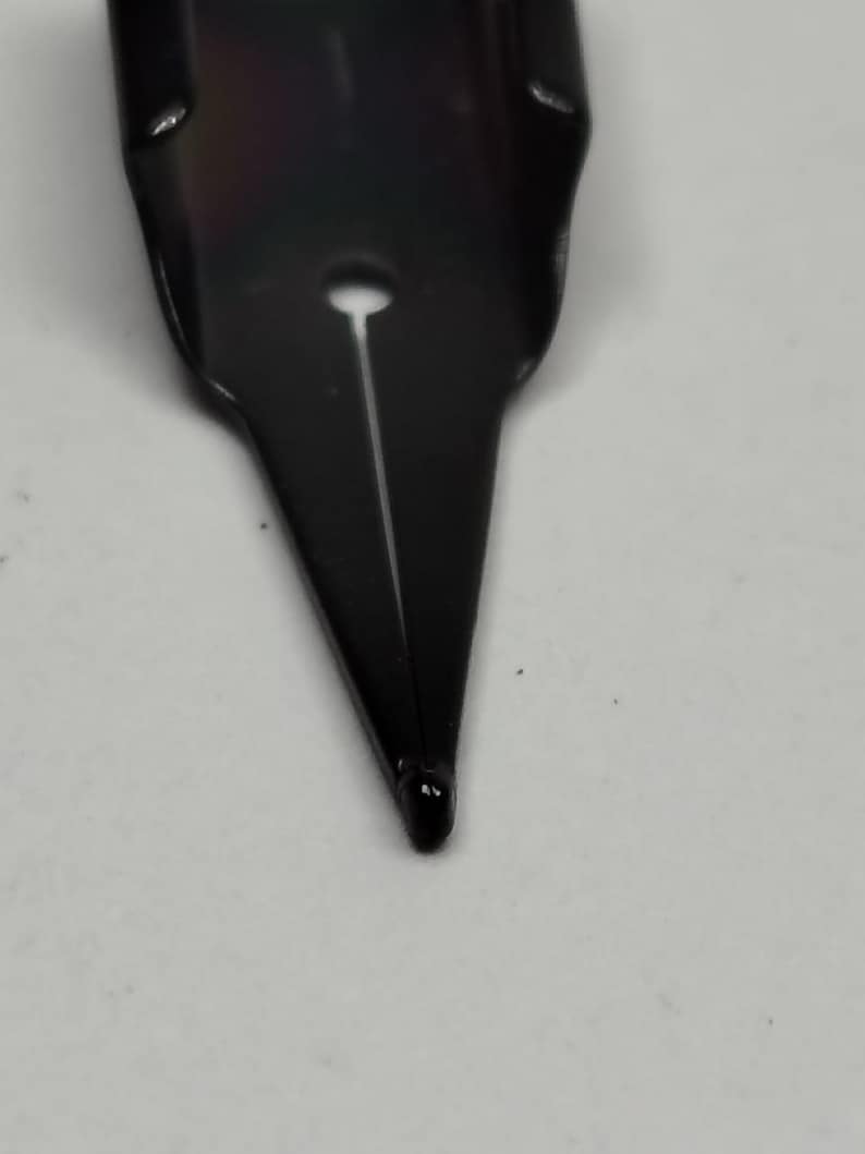 Puede incluir: Primer plano de un plum&iacute;n de pluma estilogr&aacute;fica negro. El plum&iacute;n es de metal y tiene una punta fina. Una fina ranura recorre el centro del plum&iacute;n y se ve un peque&ntilde;o agujero cerca de la parte superior. El plum&iacute;n est&aacute; sobre un fondo blanco.
