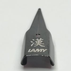 Puede incluir: Una plumilla negra con la palabra "LAMY" y un car&aacute;cter chino grabado. La plumilla tiene forma triangular y una peque&ntilde;a ranura en el centro.