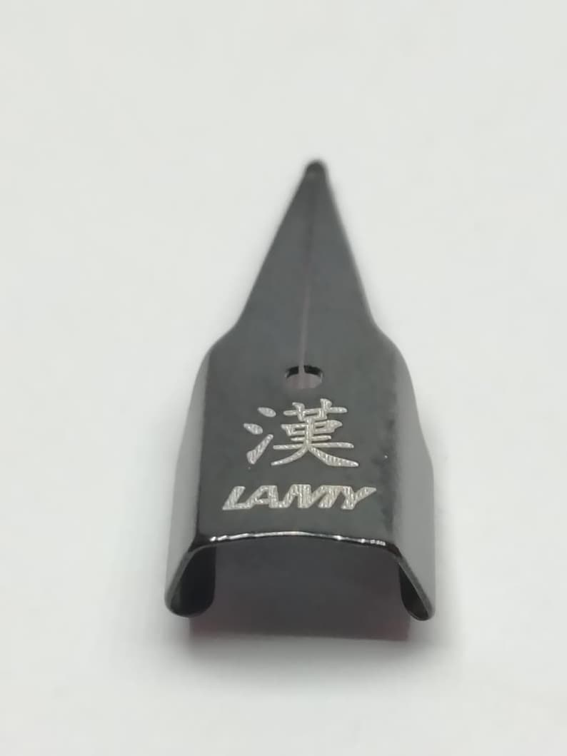 Puede incluir: Una plumilla negra con la palabra "LAMY" y un car&aacute;cter chino grabado. La plumilla tiene forma triangular y una peque&ntilde;a ranura en el centro.