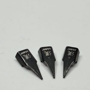 Puede incluir: Tres plumines negros Lamy para pluma estilogr&aacute;fica con caracteres japoneses y el logotipo de Lamy. Los plumines est&aacute;n orientados hacia la parte inferior derecha, con una punta afilada y puntiaguda. El fondo es gris claro liso.