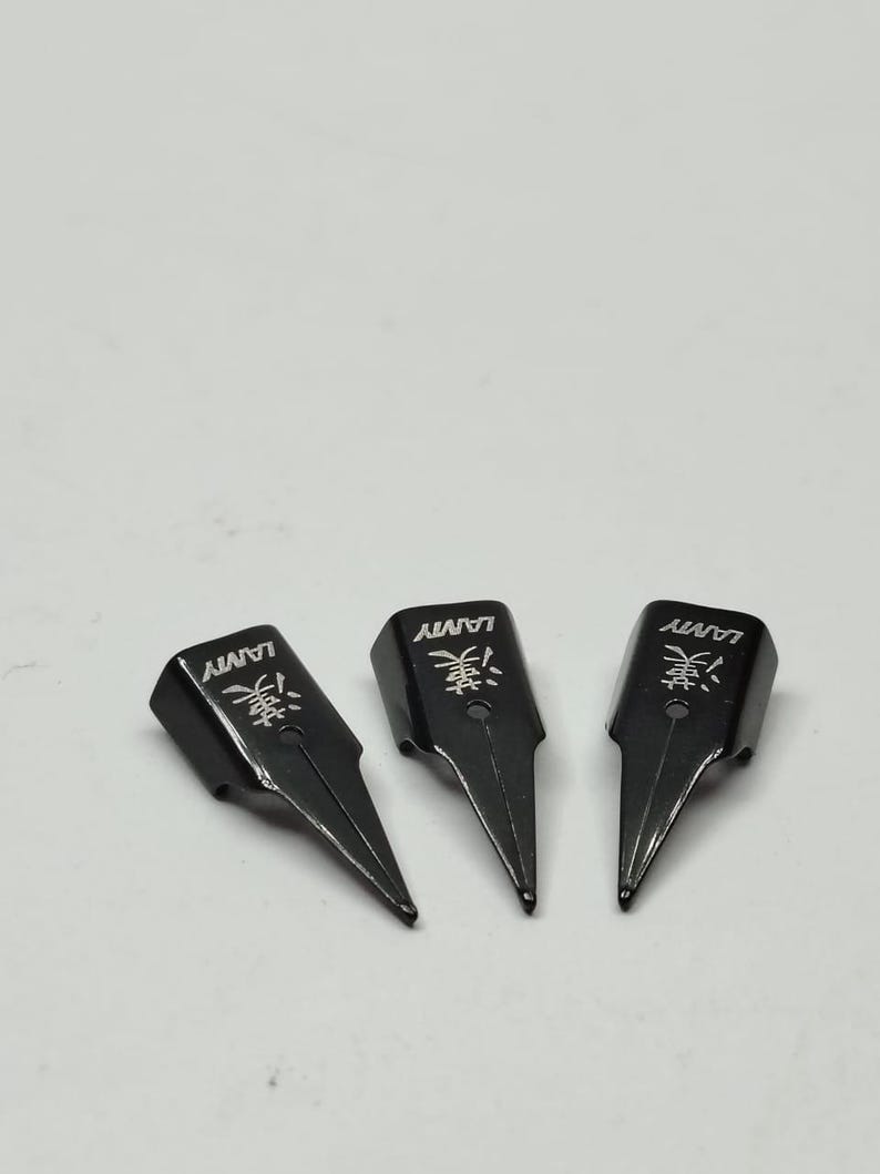 Puede incluir: Tres plumines negros Lamy para pluma estilogr&aacute;fica con caracteres japoneses y el logotipo de Lamy. Los plumines est&aacute;n orientados hacia la parte inferior derecha, con una punta afilada y puntiaguda. El fondo es gris claro liso.