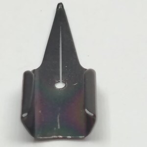 Puede incluir: Una plumilla de metal negro con punta y un peque&ntilde;o agujero. La plumilla tiene una base rectangular con lados curvos. La superficie de la plumilla tiene un ligero brillo iridiscente.