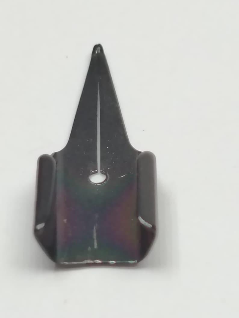 Puede incluir: Una plumilla de metal negro con punta y un peque&ntilde;o agujero. La plumilla tiene una base rectangular con lados curvos. La superficie de la plumilla tiene un ligero brillo iridiscente.