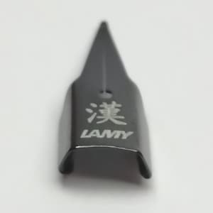 Puede incluir: Una plumilla de pluma LAMY negra con caracteres japoneses y el nombre de la marca impreso en blanco. La plumilla tiene una punta afilada y una base curva.