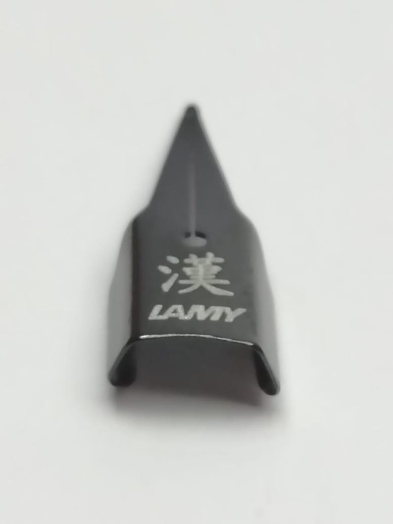 Puede incluir: Una plumilla de pluma LAMY negra con caracteres japoneses y el nombre de la marca impreso en blanco. La plumilla tiene una punta afilada y una base curva.