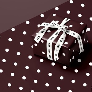 May include: A wrapped gift box with a dark burgundy polka dot pattern. The box is tied with a white ribbon featuring a repeating black logo. The gift is presented on a matching polka dot background.