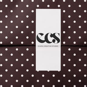 May include: A white rectangular card with the black letters "ecs" and the words "EVARA CREATIVE STUDIO" printed on it. The card is centered on a dark brown background with white polka dots.