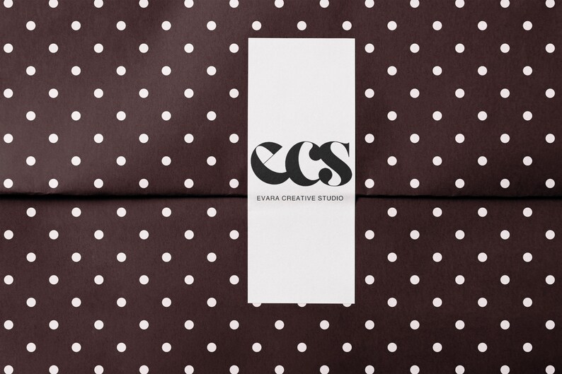 May include: A white rectangular card with the black letters "ecs" and the words "EVARA CREATIVE STUDIO" printed on it. The card is centered on a dark brown background with white polka dots.