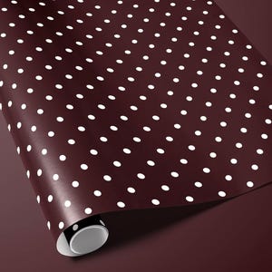 May include: A roll of maroon wrapping paper with a repeating pattern of white polka dots. The paper is partially unrolled, revealing the white interior of the roll. The background is a matching maroon color.