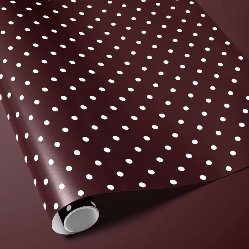 May include: A roll of maroon wrapping paper with a repeating pattern of white polka dots. The paper is partially unrolled, revealing the white interior of the roll. The background is a matching maroon color.