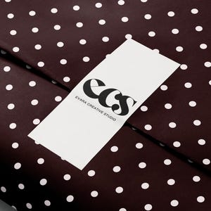 May include: A close-up of a dark brown wrapping paper with white polka dots. A white rectangular label with the text "ecs EVARA CREATIVE STUDIO" in black is placed on top of the paper. The logo features stylized black letters.