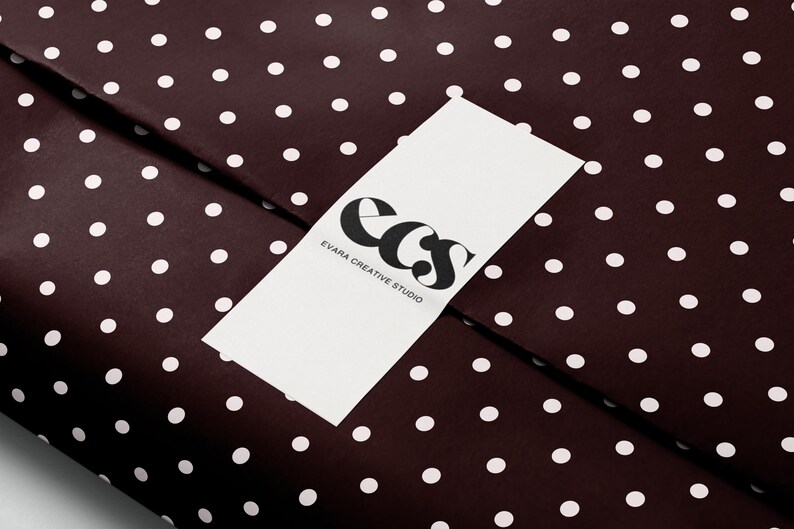 May include: A close-up of a dark brown wrapping paper with white polka dots. A white rectangular label with the text "ecs EVARA CREATIVE STUDIO" in black is placed on top of the paper. The logo features stylized black letters.