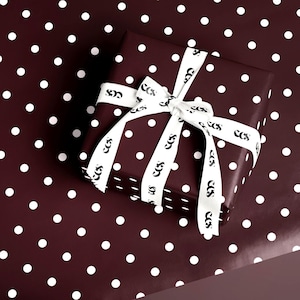 May include: A wrapped gift box with a white ribbon, set against a matching polka dot background. The box and background are a deep burgundy color with white polka dots. The white ribbon has a repeating black logo.