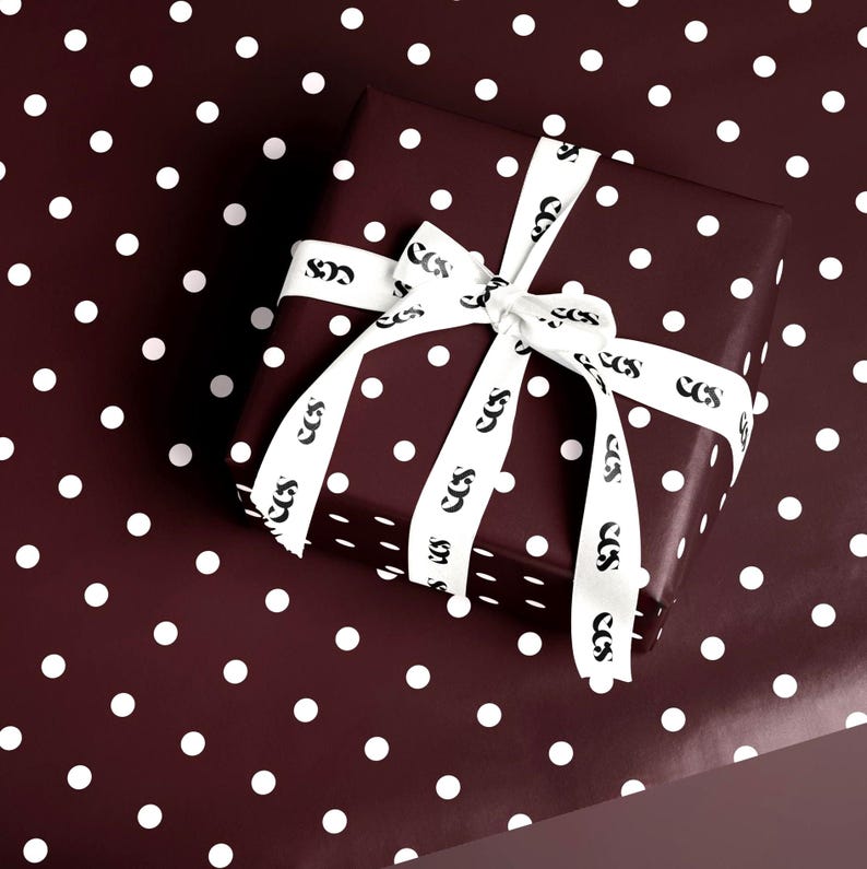 May include: A wrapped gift box with a white ribbon, set against a matching polka dot background. The box and background are a deep burgundy color with white polka dots. The white ribbon has a repeating black logo.
