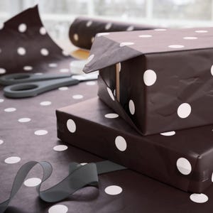 May include: Brown wrapping paper with white polka dots covers two gift boxes. A roll of matching paper and a pair of gray scissors are in the background. A gray ribbon is in the foreground, suggesting the gifts are ready to be presented.