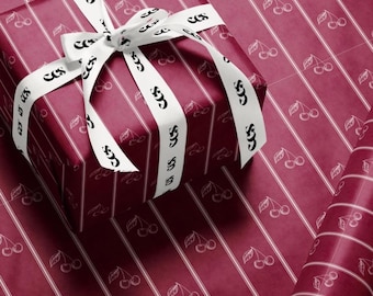May include: A square gift box wrapped in deep magenta paper featuring a white vertical striped pattern and small cherry illustrations. The box is tied with a white ribbon printed with the repeating black text "CCS". A roll of matching paper is visible.