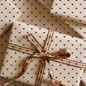 Puede incluir: Varios regalos envueltos con papel crema con un patrón de pequeños corazones rojos. Los regalos están atados con una cinta decorativa con un diseño floral y un borde marrón. Los regalos están dispuestos sobre una superficie crema.