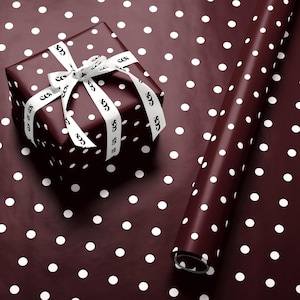 May include: A wrapped gift box and a roll of wrapping paper. Both are dark burgundy with white polka dots. The gift is tied with a white ribbon printed with a repeating design. The wrapping paper is rolled up.