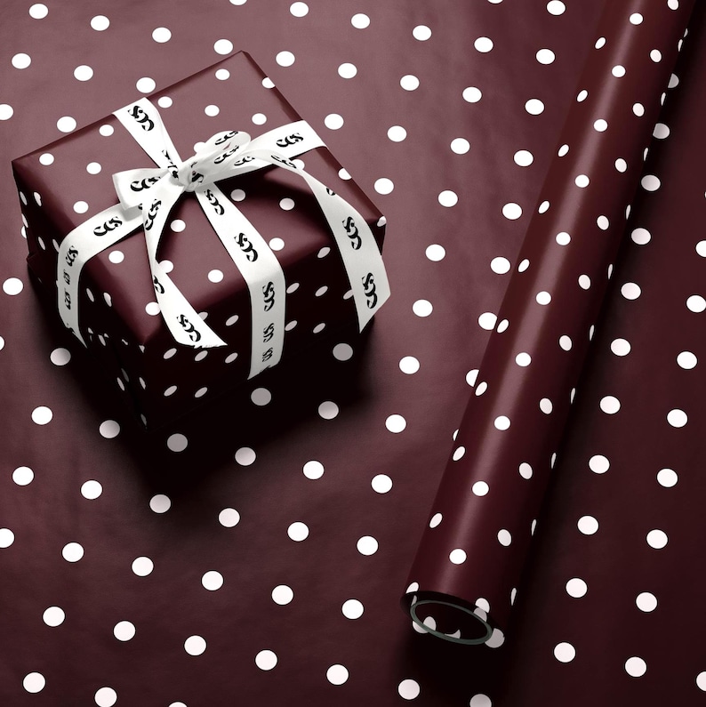 May include: A wrapped gift box and a roll of wrapping paper. Both are dark burgundy with white polka dots. The gift is tied with a white ribbon printed with a repeating design. The wrapping paper is rolled up.