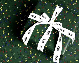 May include: A rectangular gift box wrapped in dark green paper featuring a repeating pattern of black tangled wires and multi-colored Christmas light bulbs in red, blue, and yellow. A white ribbon with the black text CCS is tied in a bow on top.
