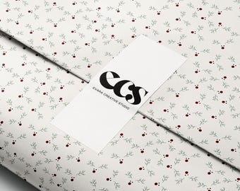 Op de afbeelding: Cadeaupapier met een herhalend patroon van groene takjes en rode bessen op een witte achtergrond. Een wit label met de tekst "ECS EVARA CREATIVE STUDIO" is erop geplaatst.
