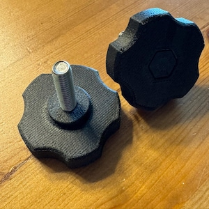 May include: Two black, star-shaped knobs with a silver bolt extending from the center. The knobs have a textured surface and a hexagonal indentation in the center. One knob is shown with the bolt attached, and the other is separate.