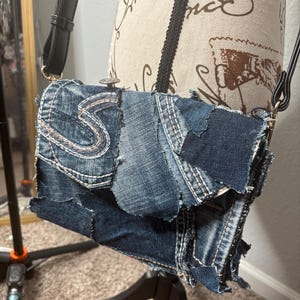 May include: A denim crossbody bag with a patchwork design. The bag has a flap closure and a black adjustable strap. The denim is a mix of light and dark washes, with visible stitching and frayed edges. The bag is displayed on a mannequin.