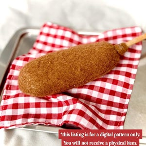 May include: A brown, textured corndog on a wooden stick, resting on a red and white checkered napkin. The napkin is placed on a silver tray. Text at the bottom reads: "*this listing is for a digital pattern only. You will not receive a physical item."