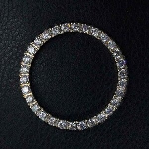 May include: A gold-toned, circular bracelet with numerous clear, sparkling gemstones set closely together. The bracelet is displayed against a dark, textured background, highlighting the brilliance of the stones. The design suggests a luxury jewelry item.