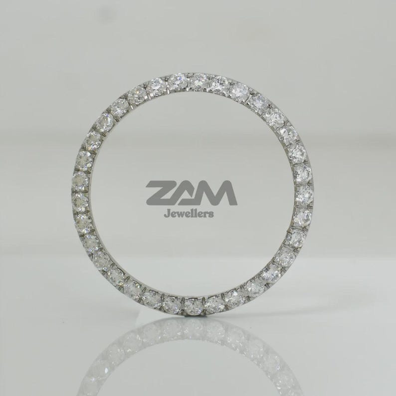 K&ouml;nnte beinhalten: Ein kreisf&ouml;rmiges Schmuckst&uuml;ck mit einem Ring aus kleinen, klaren Edelsteinen. Das St&uuml;ck ist vor einem wei&szlig;en Hintergrund platziert, mit dem Text "ZAM Jewellers" in Grau.