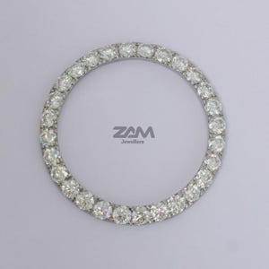May include: Circular jewelry piece with numerous clear gemstones set in a silver-colored band. The gemstones are evenly spaced around the circle. The text "ZAM Jewellers" is visible in the center.