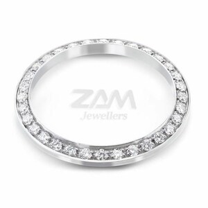 May include: A silver-toned watch bezel with a ring of small, clear gemstones. The bezel is circular and has the text "ZAM Jewellers" in the center. The gemstones are evenly spaced around the outer edge of the bezel.
