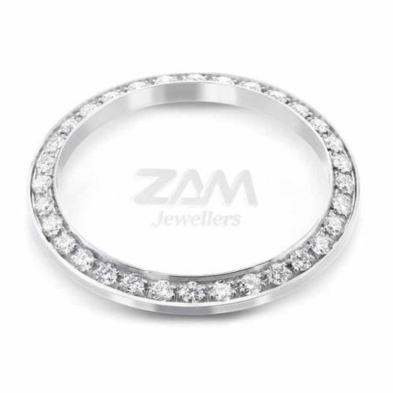 May include: A silver-toned watch bezel with a ring of small, clear gemstones. The bezel is circular and has the text "ZAM Jewellers" in the center. The gemstones are evenly spaced around the outer edge of the bezel.