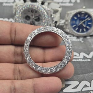 May include: A silver watch bezel set with a ring of clear, round gemstones. The bezel is held in a hand, with two diamond-encrusted watches in the background. The watches have white and blue faces.