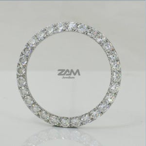 May include: A circular, silver-toned jewelry piece, encrusted with numerous clear, sparkling gemstones. The gemstones are evenly spaced around the entire circle. The brand name "ZAM Jewellers" is visible in the center. The piece is set against a light background.