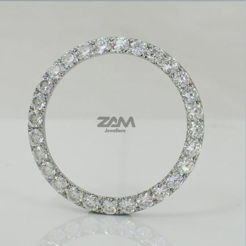 May include: A circular, silver-toned jewelry piece, encrusted with numerous clear, sparkling gemstones. The gemstones are evenly spaced around the entire circle. The brand name "ZAM Jewellers" is visible in the center. The piece is set against a light background.