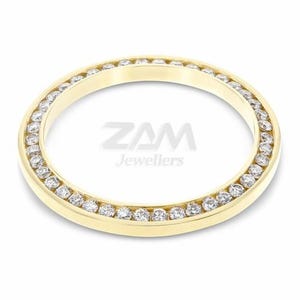 May include: A gold-colored ring with a circular design, featuring a row of small, clear gemstones set around the outer edge. The text "ZAM Jewellers" is visible in the center.