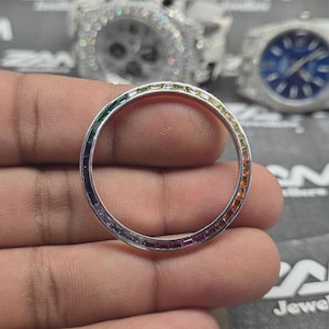 以下が含まれることがあります： 長方形の宝石が付いたレインボーカラーのベゼルリング。リングは手で持たれており、背景にはダイヤモンドをちりばめた2つの時計があります。宝石は緑から赤へと変化します。