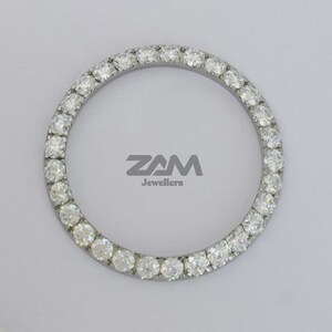 K&ouml;nnte beinhalten: Ein kreisf&ouml;rmiges Schmuckst&uuml;ck mit einem Ring aus kleinen, klaren Edelsteinen. Die Edelsteine sind in einem silberfarbenen Metall gefasst. Der Text "ZAM Jewellers" ist grau gedruckt.