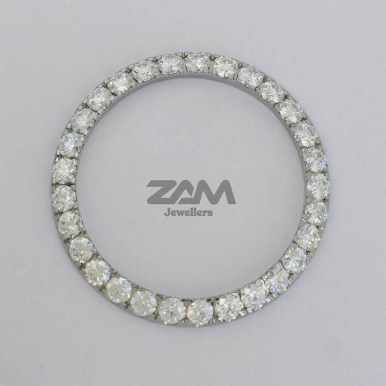 K&ouml;nnte beinhalten: Ein kreisf&ouml;rmiges Schmuckst&uuml;ck mit einem Ring aus kleinen, klaren Edelsteinen. Die Edelsteine sind in einem silberfarbenen Metall gefasst. Der Text "ZAM Jewellers" ist grau gedruckt.