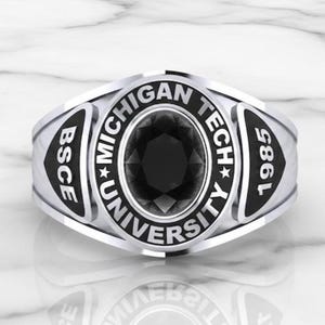 Pode incluir: Um anel prateado com uma pedra preta em forma oval no centro. O anel apresenta as palavras "MICHIGAN TECH UNIVERSITY", "BSCE" e "1985" em letras pretas. O anel tem um acabamento polido.