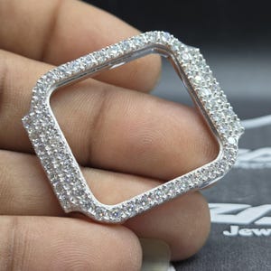 May include: A silver-coloured, square watch case, studded with many small, clear, sparkling gemstones. The case has a decorative, slightly curved design on the corners. The watch case is held in a hand against a dark background.