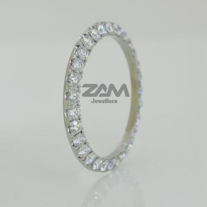 K&ouml;nnte beinhalten: Ein silberner Ring mit einer durchgehenden Reihe klarer Edelsteine. Der Ring ist vor einem hellen Hintergrund platziert. Der Text "ZAM Jewellers" ist auf dem Ring zu sehen.
