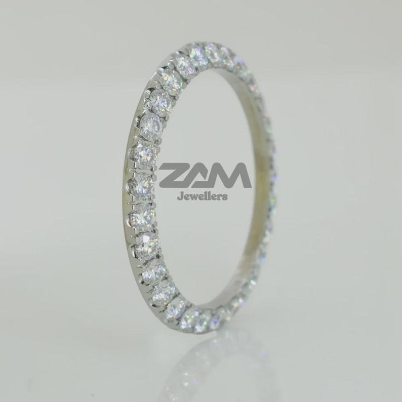 K&ouml;nnte beinhalten: Ein silberner Ring mit einer durchgehenden Reihe klarer Edelsteine. Der Ring ist vor einem hellen Hintergrund platziert. Der Text "ZAM Jewellers" ist auf dem Ring zu sehen.