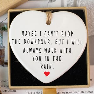 Könnte beinhalten: Eine weiße, herzförmige Keramikplakette mit dem Text "MAYBE I CAN'T STOP THE DOWNPOUR, BUT I WILL ALWAYS WALK WITH YOU IN THE RAIN." Ein kleines rotes Herz befindet sich unten. Die Plakette befindet sich in einer Geschenkbox.