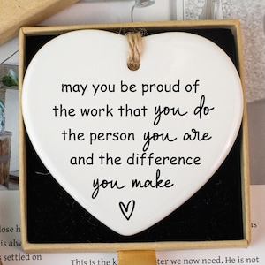 Könnte beinhalten: Eine weiße herzförmige Keramikplakette mit einer Botschaft, präsentiert in einer Geschenkbox. Die Plakette hat eine Juteschnur zum Aufhängen und eine Botschaft mit der Aufschrift "may you be proud of the work that you do..." Die Box ist braun mit einem "Hand made"-Etikett.